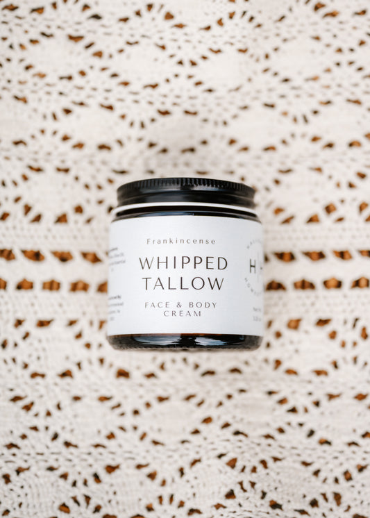 A jar of Frankincense Whipped Tallow Face & Body Cream, featuring a white label and black lid, placed on a textured cream crochet blanket.
