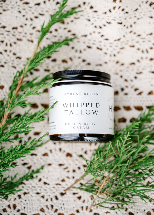 A jar of Forest Blend Whipped Tallow Face & Body Cream, styled with green pine branches on a textured cream crochet blanket.